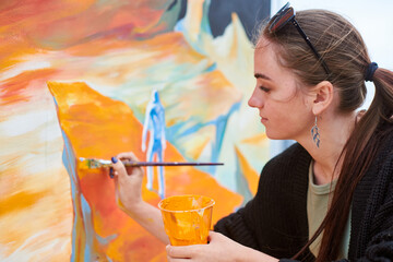 Attractive female painter passionately draws picture with paintbrush for outdoor street exhibition,...