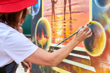 Female painter in red hat draws picture with paintbrush on canvas for outdoor street exhibition, close up side view of female artist apply brushstrokes to canvas, symphony of art creativity