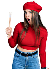 Young brunette teenager holding paintbrushes wearing beret scared and amazed with open mouth for...