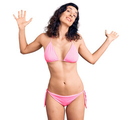 Young beautiful hispanic woman wearing bikini clueless and confused expression with arms and hands...
