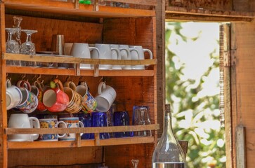 Wooden cupboard with glasses and cups on it in a cabin, green wilderness visible through the door