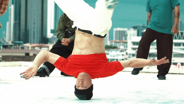 Professional break dancer doing head spin at roof top with city view. Skilled caucasian hipster practicing break dancing with multicultural friends encourage behind. Outdoor sport 2024. hiphop.