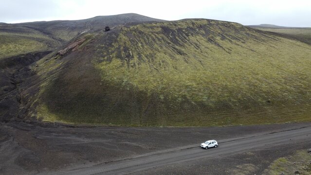 Drone shot of a car going by a green moss-covered volcanic mountain and black lava field, Iceland