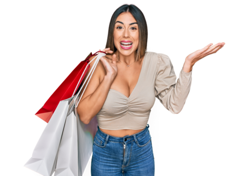 Young hispanic woman holding shopping bags celebrating victory with happy smile and winner expression with raised hands