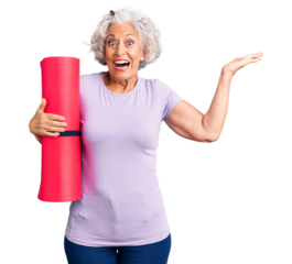 Senior grey-haired woman holding yoga mat celebrating victory with happy smile and winner expression with raised hands