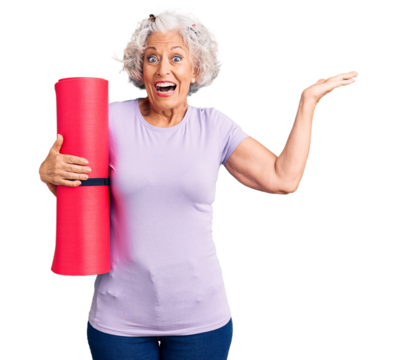 Senior grey-haired woman holding yoga mat celebrating victory with happy smile and winner expression with raised hands
