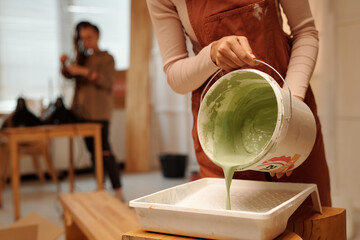 Cropped shot of young woman in overalls bending over bench and pouring green paint in plastic tray...