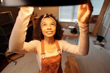 Young African American businesswoman in workwear hanging new lamp on ceiling of cafe while making...