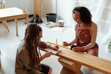 Two young creative female designers with pencils sitting on the floor by wooden bench while taking measures and making notes - 775042556