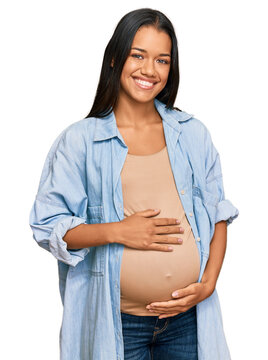 Fototapeta Beautiful hispanic woman expecting a baby showing pregnant belly looking positive and happy standing and smiling with a confident smile showing teeth