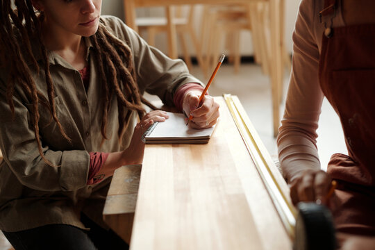 Young woman in grey shirt holding pencil over blank page of copybook while sitting by wooden bench in front of female colleague
