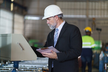 A man in a suit is looking at a computer screen while holding a clipboard to check product quality in the factory.
