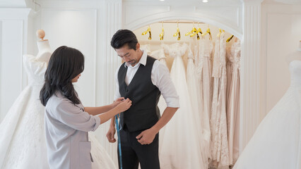 A man and woman are standing in front of a rack of wedding dresses. The owner of the clothing store measures the groom's body to make a suit.