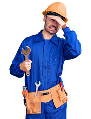 Young hispanic man wearing electrician uniform holding wrench stressed and frustrated with hand on head, surprised and angry face