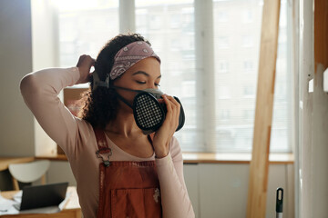Young African American woman putting on protective respirator to prevent herself from toxic smell of paint before doing renovation work - 775046997
