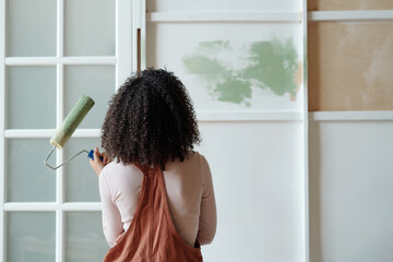 Rear view of young African American female owner of cafe with paintroller in hand standing in front of wall while painting it in green color - 775049348