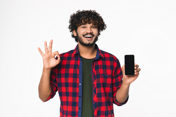 Positive young Hindi man showing mobile phone screen and okay gesture isolated over white...