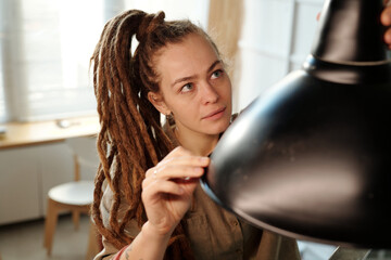 Young female owner of cafe or restaurant with dreadlocks looking at new lamp while hanging it over table during renovation works - 775050538
