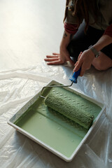 High angle of hand of young female worker sitting on knees on the floor covered by cellophane and putting paintroller in tray with green paint - 775050731