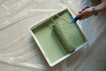 Top view of white plastic tray with green paint standing on the floor covered with cellophane and hand of woman holding paintroller - 775050912