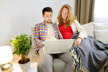 A couple chooses something online using their modern laptop while sitting on the couch at home. A young family is expecting a child.