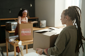 Young businesswoman with dreadlocks carrying packed cardboard box while moving towards her colleague unpacking fragile items