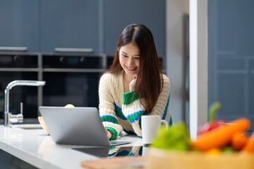 Happy woman using laptop - young Asian woman studying while sitting at  table online learning by laptop computer at home