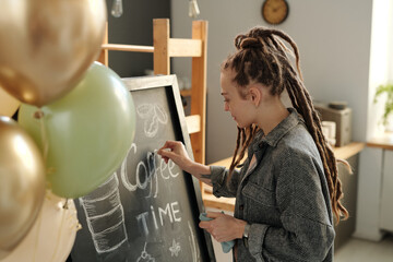 Young businesswoman with dreadlocks standing in front of blackboard and drawing advert of new cafe...