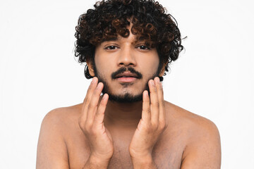 Handsome shirtless Hindu man looking at the mirror shaving bear isolated over white background. Young Indian boy removing facial hair, touching his cheeks