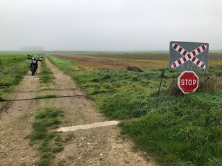 Railway track crossing a rural road in Hauts-de-France. A motorbike waits to pass. - 775058901