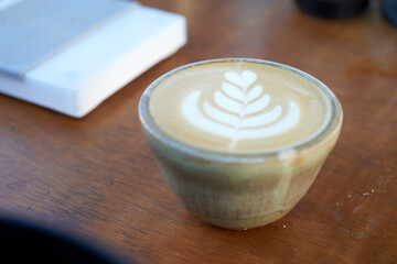 A beautifully crafted cappuccino with a detailed latte art design sits on a wooden table, accompanied by a closed book and other blurred objects in the background