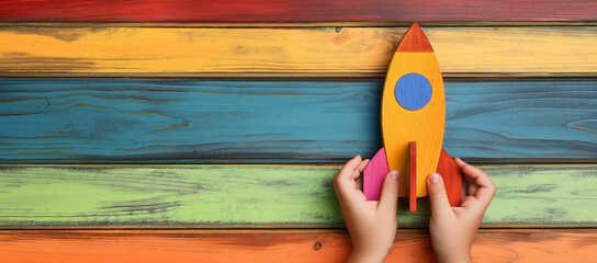 Child's Hands Holding Colorful Wooden Rocket Toy Against Vibrant Multicolored Plank Background