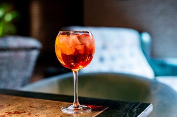 An orange cocktail in a wine glass on a rustic table, with a blurred lounge setting in the background. The vibrant drink features ice cubes and may be an Aperol Spritz.