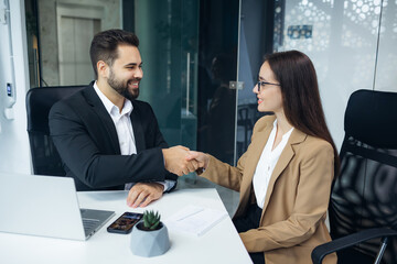 Caucasian man and woman in formal wear making hands shaking after serious conversation at modern office. Two partners sitting together at desk and having successful deal about new business project. - 775083554