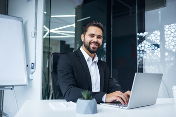 Modern Office Portrait of Stylish Hispanic Businessman Work on Laptop. makes Data Analysis, Looks at Camera and Smiles. Entrepreneur Works on e-Commerce Startup Project