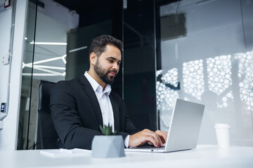 thoughtful male businessman working on laptop computer at a modern office desk. Confident Focused pensive business man employee in suit indoor.