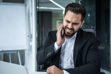 Toothache in the workplace. A young man, a businessman, an employee, has a teeth pain at work. He is sitting at a desk in the office, holding his cheek