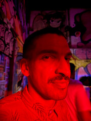 A man with a mustache and red shirt is standing in front of a wall with graffiti