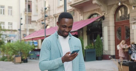 Happy african american man using his mobile phone in stylish clothes walking near cozy coffee shop in old city part