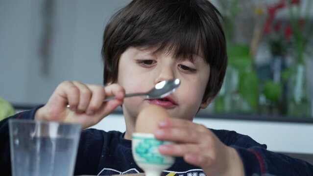 Child cracking open egg with spoon at breakfast table. One concentrated small boy opening soft-boiled egg with spoon