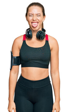 Beautiful blonde woman wearing gym clothes and using headphones sticking tongue out happy with funny expression. emotion concept.