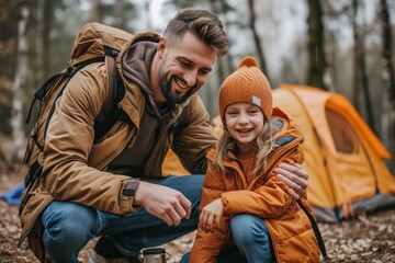 Smiling father, crouched next to daughter in orange jacket, shares a joyful moment beside tent in woods. Happy man kneels with laughing child, clothed in warm gear, during forest camping adventure. - Powered by Adobe
