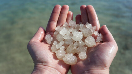 A pair of open hands holding clear, glistening Dead Sea salt crystals against a backdrop of its iconic, mineral-rich waters. Ideal for wellness and natural beauty themes