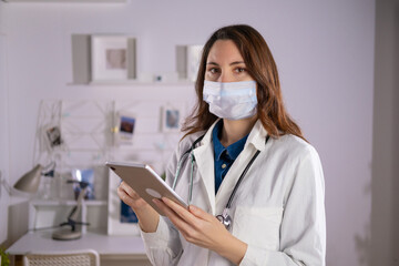 A female doctor in a mask and white uniform stands with a tablet in her hands and looks at the camera