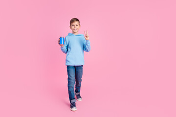Full length portrait of positive charming boy walk hold soda can show v-sign empty space isolated on pink color background