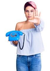 Young beautiful woman with pink hair holding vintage telephone with open hand doing stop sign with serious and confident expression, defense gesture