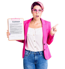 Young beautiful woman with pink hair holding clipboard with contract document surprised pointing with finger to the side, open mouth amazed expression.