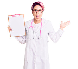 Young beautiful woman with pink hair wearing doctor stethoscope holding clipboard with medical report celebrating victory with happy smile and winner expression with raised hands