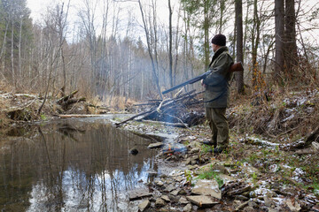 A hunter stands next to a campfire on the bank of a forest river