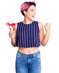 Young beautiful woman with pink hair holding graduate degree diploma pointing thumb up to the side smiling happy with open mouth